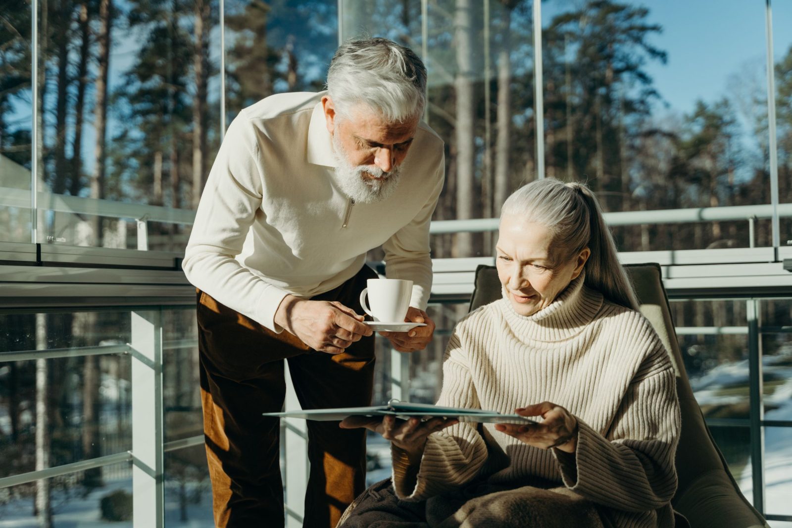 Ein Mann bringt einer Frau im Sessel eine Tasse Kaffee, vor der Glasfront einer sonnigen Veranda