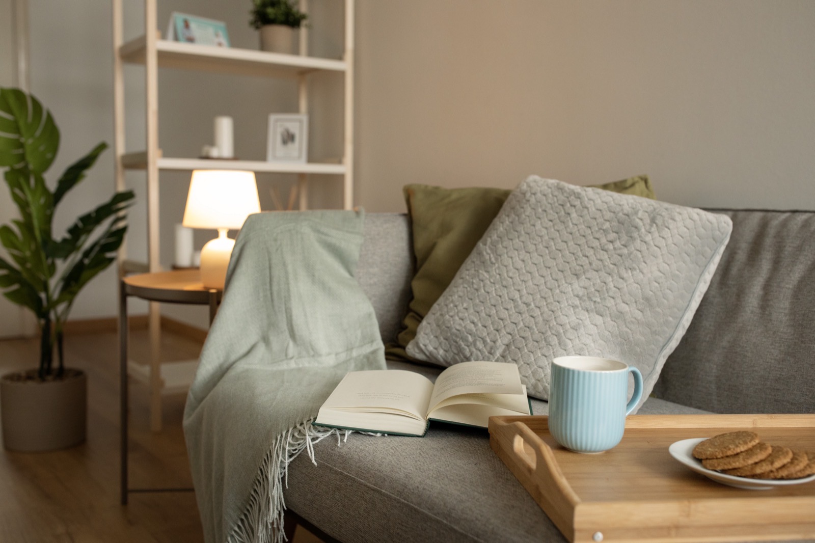 Ein Sofa mit Decke, aufgeschlagenem Buch und einem Tablett mit Tasse und Keksen in einem von einer Lampe beleuchteten Wohnzimmer